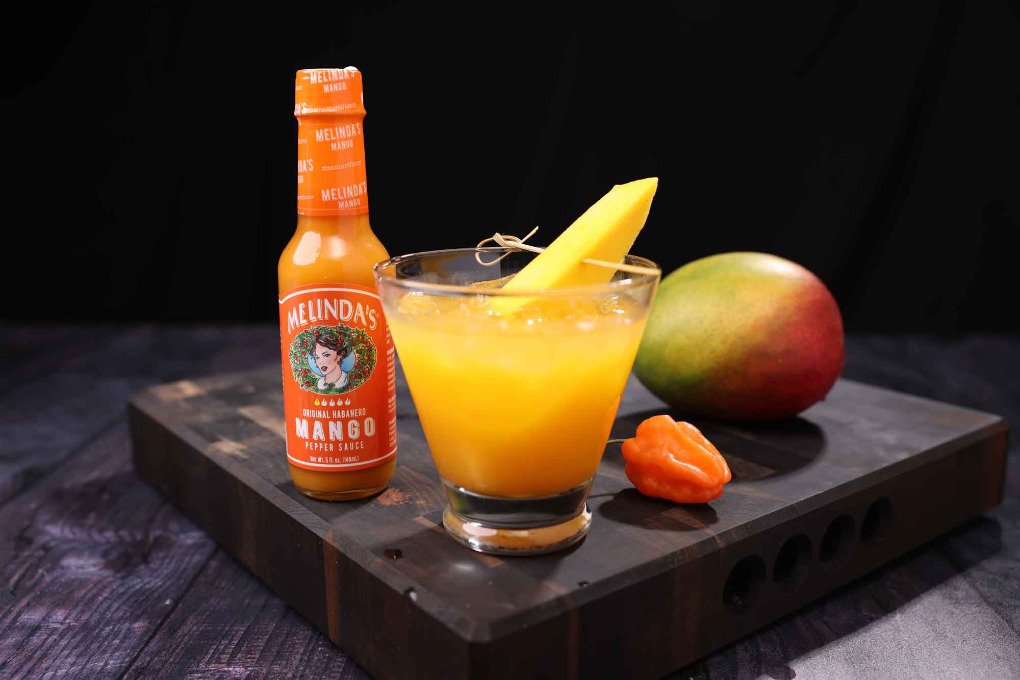 Mango rum cocktail with orange garnish next to Melinda's mango hot sauce bottle and fresh mango on wooden board