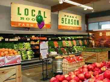 Grocery store produce section with "LOCAL ROOTS FARM" and "IN SEASON" signs above displays of fresh fruits and vegetables.
