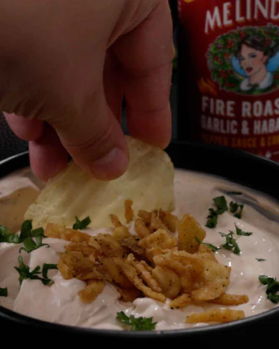 Hand dipping chip into creamy french onion dip with crispy onions and herbs on white plate