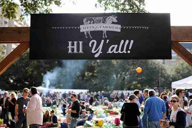 Large outdoor farmers market event with "Hi Y'all!" welcome sign and crowds of people browsing vendor booths under trees