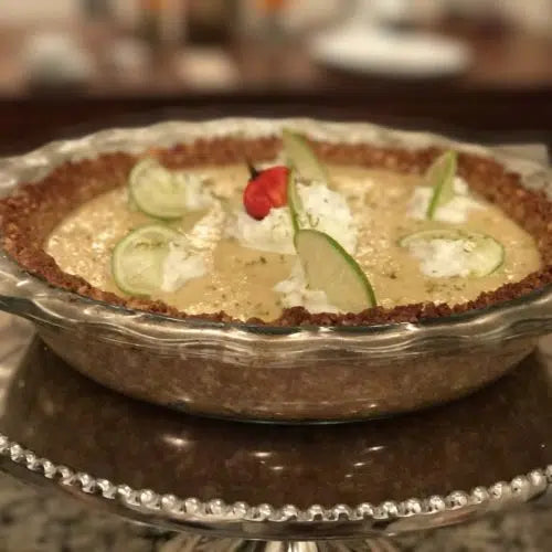 Key lime pie in glass pie dish topped with lime slices, whipped cream dollops, and red garnish on decorative plate
