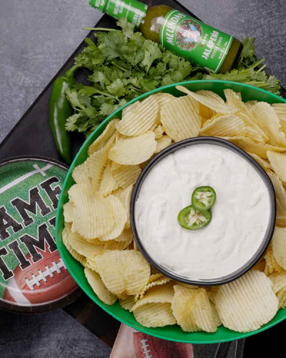 Potato chips arranged around a bowl of white sour cream dip topped with jalapeño slices, with beer and football nearby.