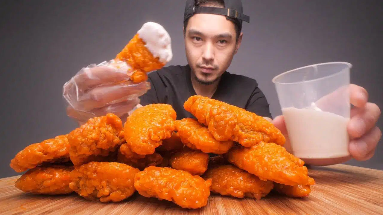 Man holding spicy orange buffalo chicken tender and glass of milk, with large pile of buffalo tenders on wooden table