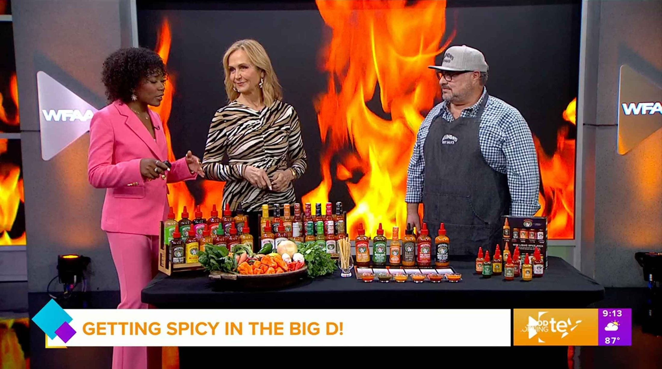 Three people on TV set discussing hot sauce products with flame backdrop and 