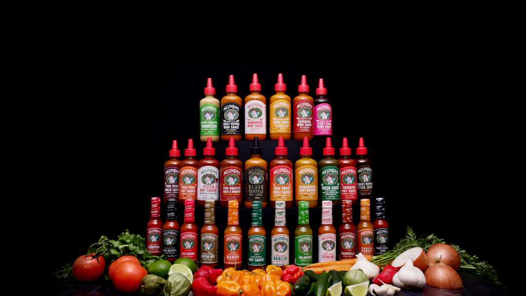 Collection of Melinda's hot sauce bottles in various flavors arranged in rows with fresh vegetables and ingredients...