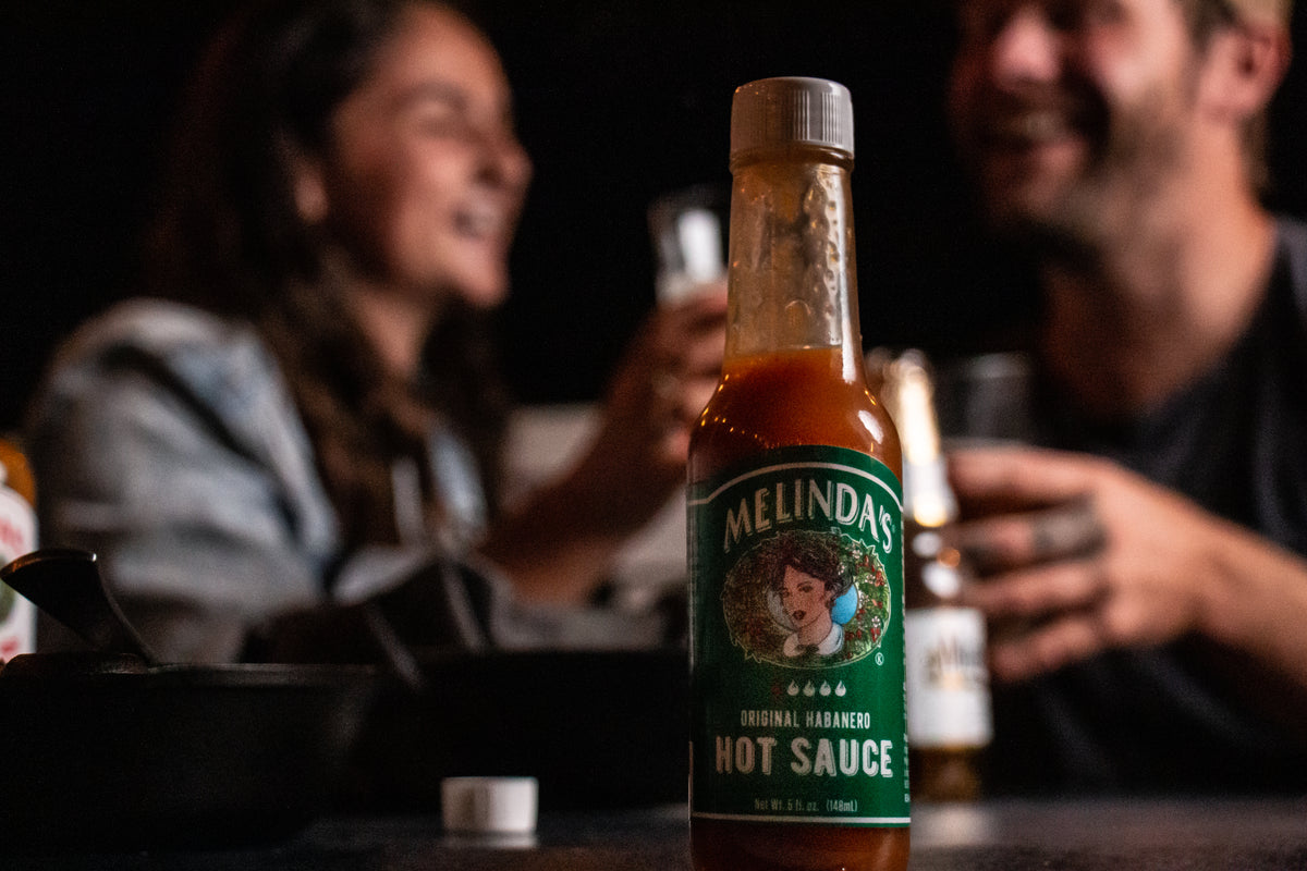 Melinda's Original Habanero Hot Sauce bottle with green label on table with blurred people socializing in background