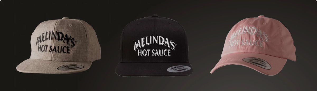 Three baseball caps in tan, black, and pink colors, each featuring "Melinda's Hot Sauce" logo embroidered on the front.