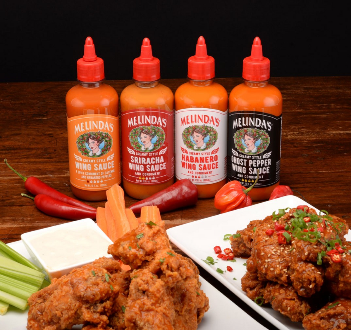 Four bottles of Melinda's wing sauce varieties with fried chicken wings, fresh peppers, and celery on wooden surface