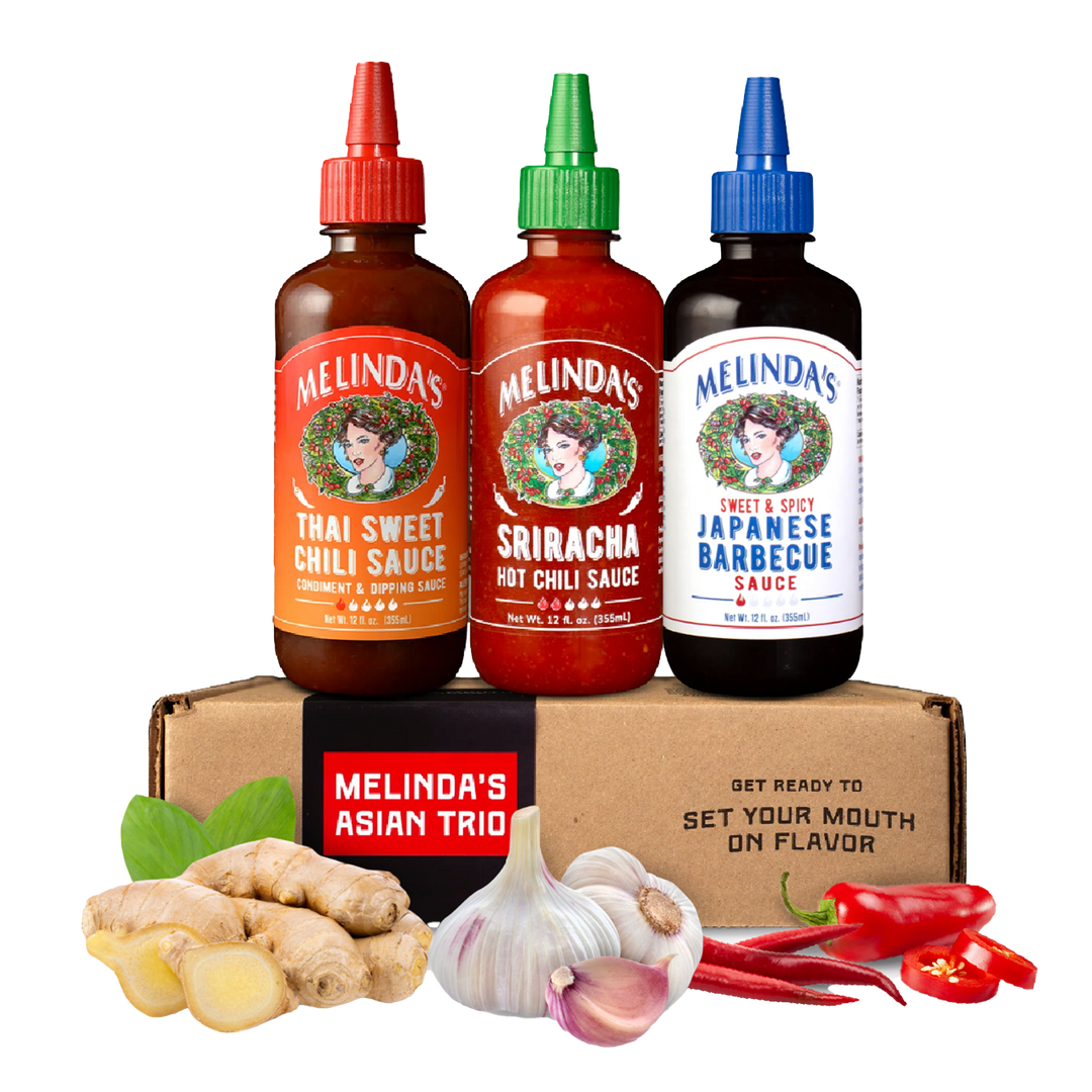 Three Melinda's sauce bottles (Thai Sweet Chili, Sriracha, Japanese Barbecue) on cardboard box with fresh ginger, garlic,...