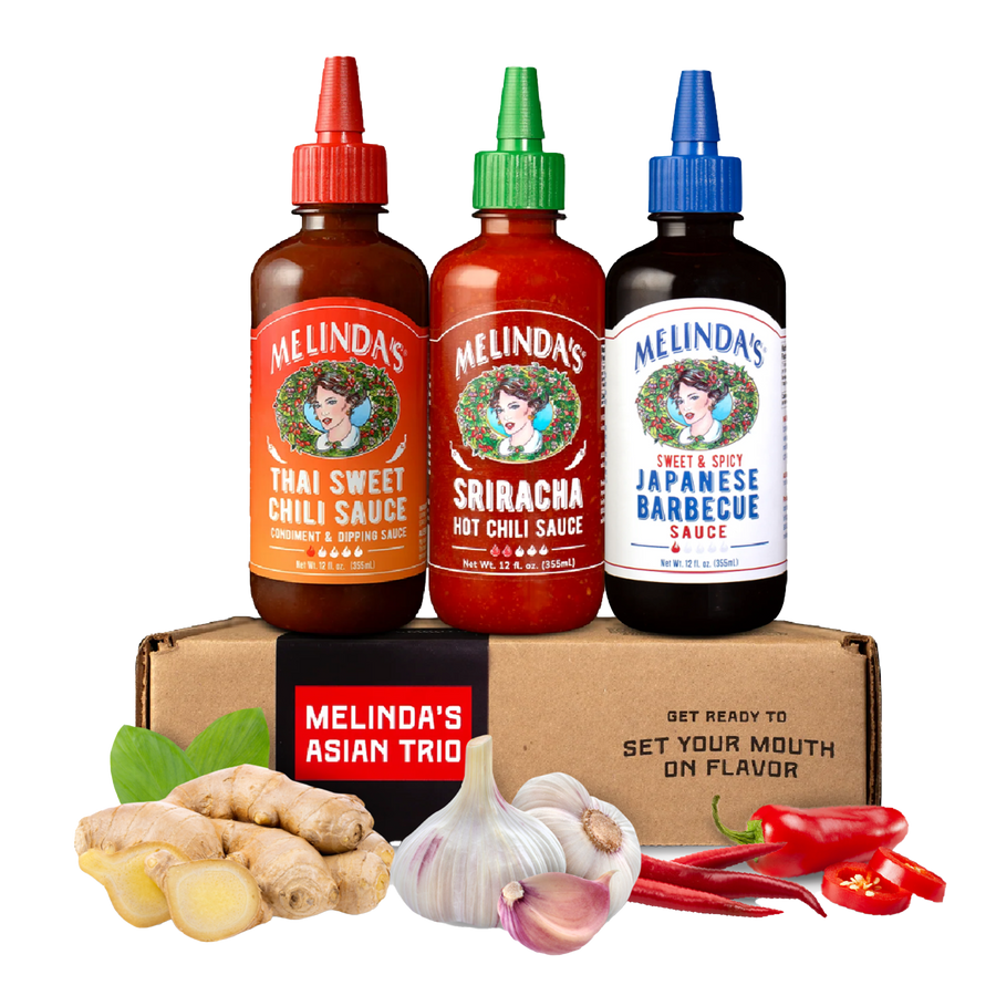 Three Melinda's sauce bottles (Thai Sweet Chili, Sriracha, Japanese Barbecue) on cardboard box with fresh ginger, garlic,...