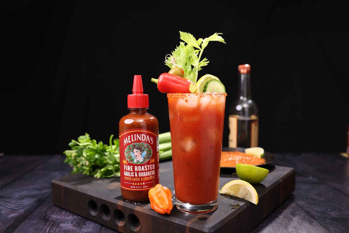 Bloody Mary cocktail with celery garnish next to Melinda's Fire Roasted Garlic & Habanero hot sauce bottle on wooden board