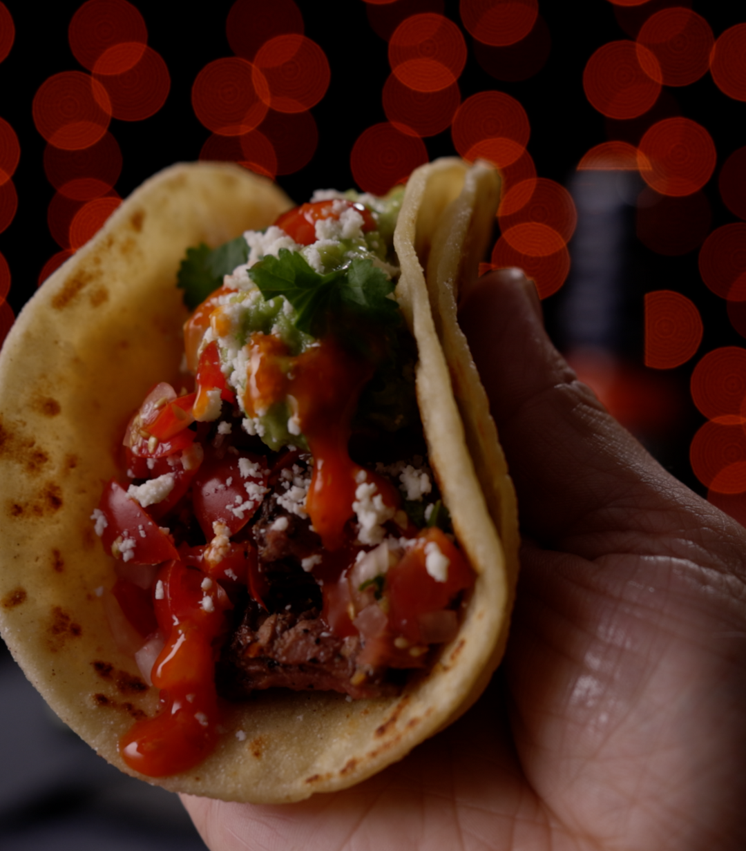 Hand holding a taco filled with meat, red peppers, white cheese, and cilantro against blurred red lights background