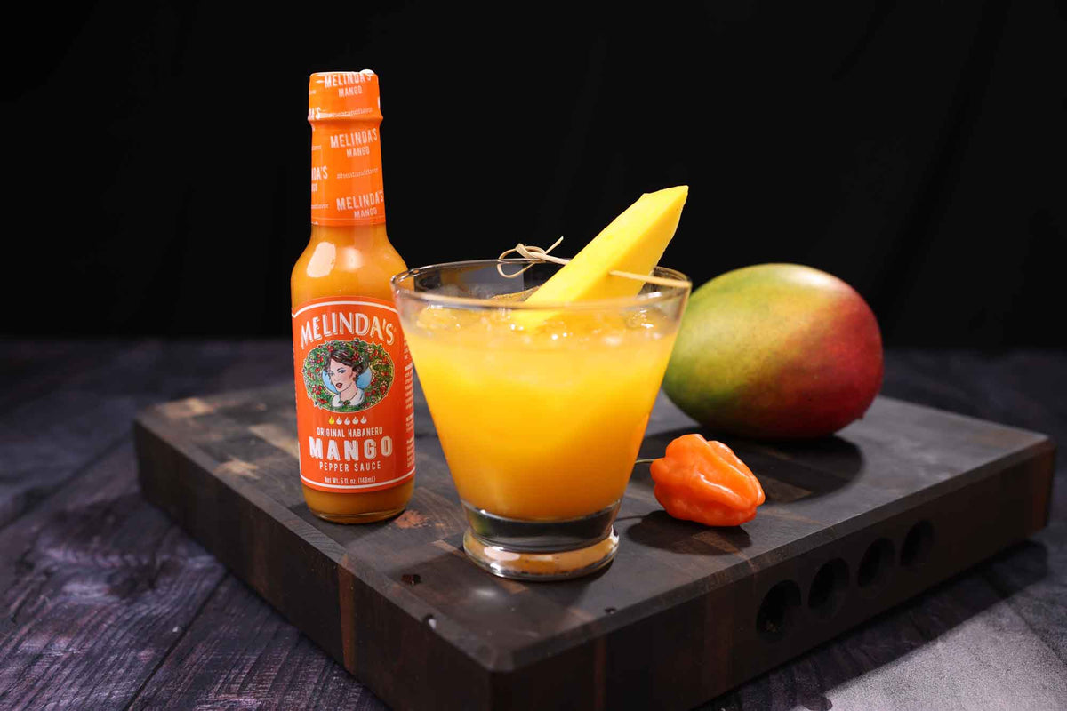 Orange Melinda's Mango hot sauce bottle next to yellow cocktail garnished with mango slice, whole mango and habanero...