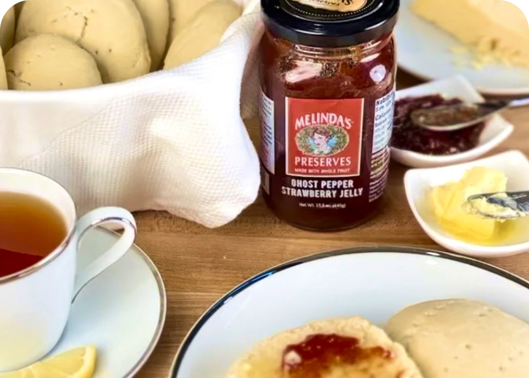 Jar of Melinda's Ghost Pepper Strawberry Jelly on breakfast table with tea cup, buttered scones, and fresh fruit