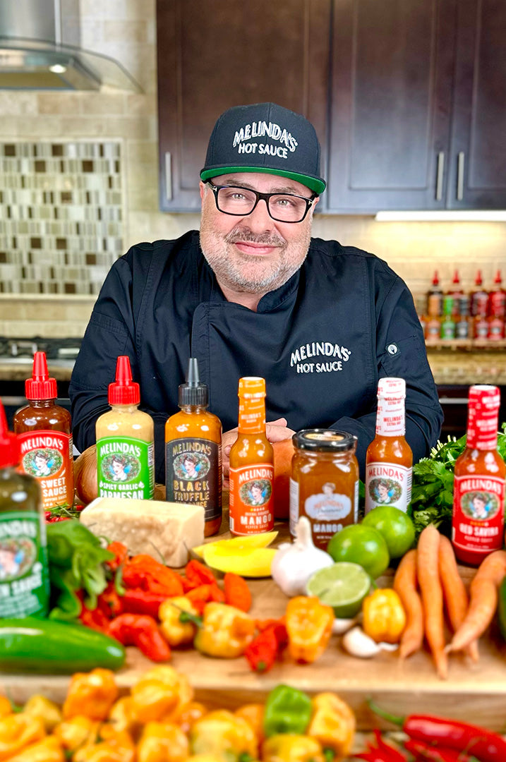 Chef in Melinda's Hot Sauce hat and apron smiling behind kitchen counter with various hot sauce bottles and fresh vegetables
