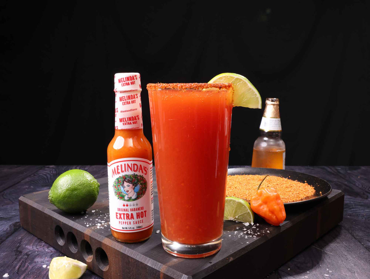 Spicy red cocktail with lime garnish next to Melinda's Extra Hot sauce bottle on wooden serving board with lime and peppers