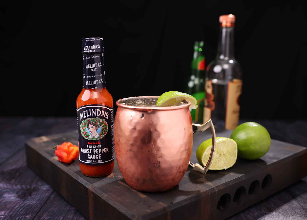 Copper Moscow mule mug with lime garnish next to Melinda's Ghost Pepper hot sauce bottle on wooden cutting board