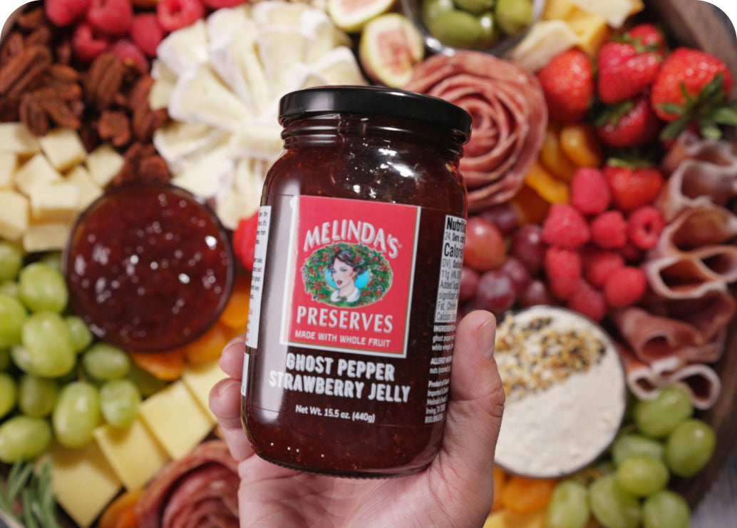 Hand holding jar of Melinda's Ghost Pepper Strawberry Jelly against backdrop of charcuterie board with cheeses, meats,...