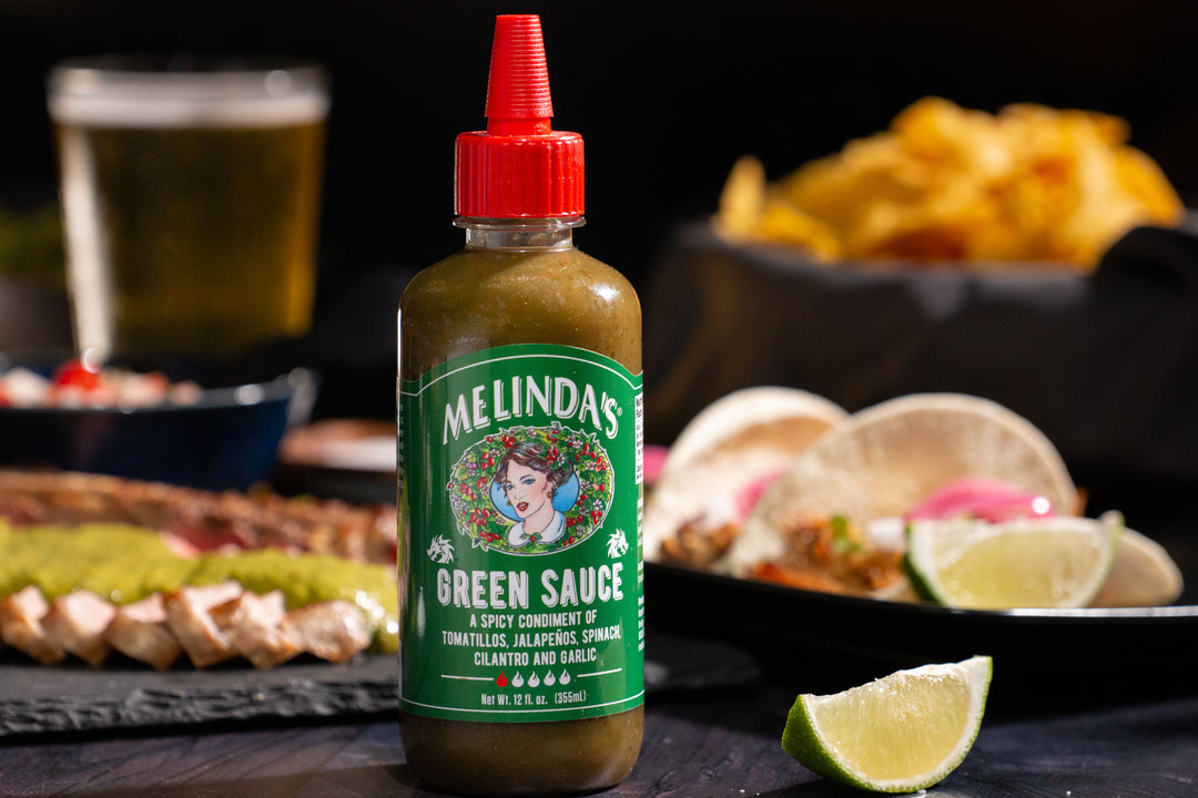 Melinda's Green Sauce bottle with red cap on dark surface, surrounded by lime wedges, tacos, chips, and beer in background