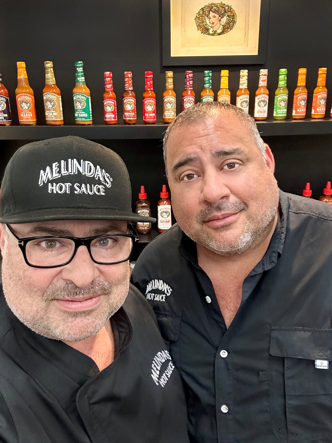 Two men in black Melinda's Hot Sauce shirts pose together in front of a colorful display of hot sauce bottles on shelves.
