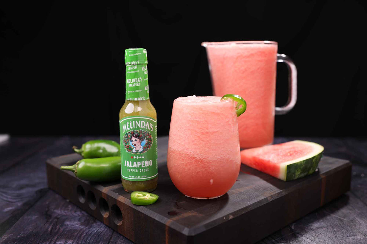 Spicy watermelon drink in glass with jalapeño garnish, pitcher, Melinda's jalapeño sauce bottle, and fresh ingredients