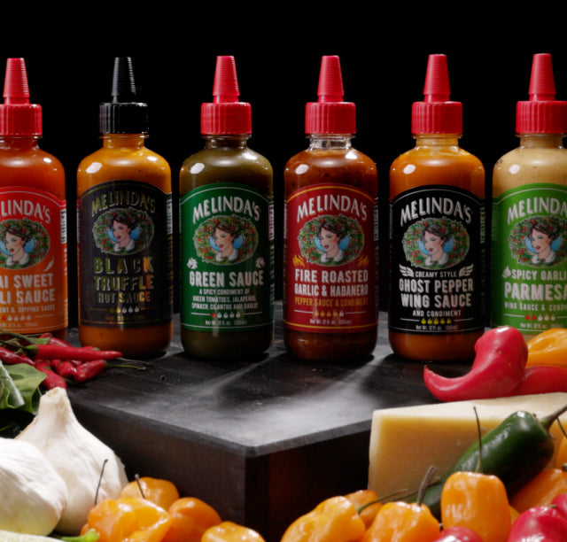 Six Melinda's hot sauce bottles with colorful labels arranged on dark surface with fresh peppers and vegetables