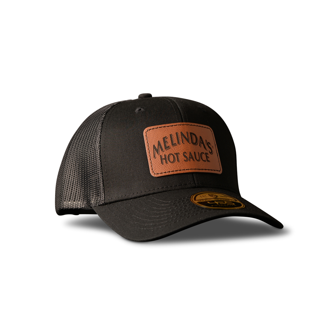 Black trucker hat with brown leather patch displaying "MELINDA'S HOT SAUCE" text on white background