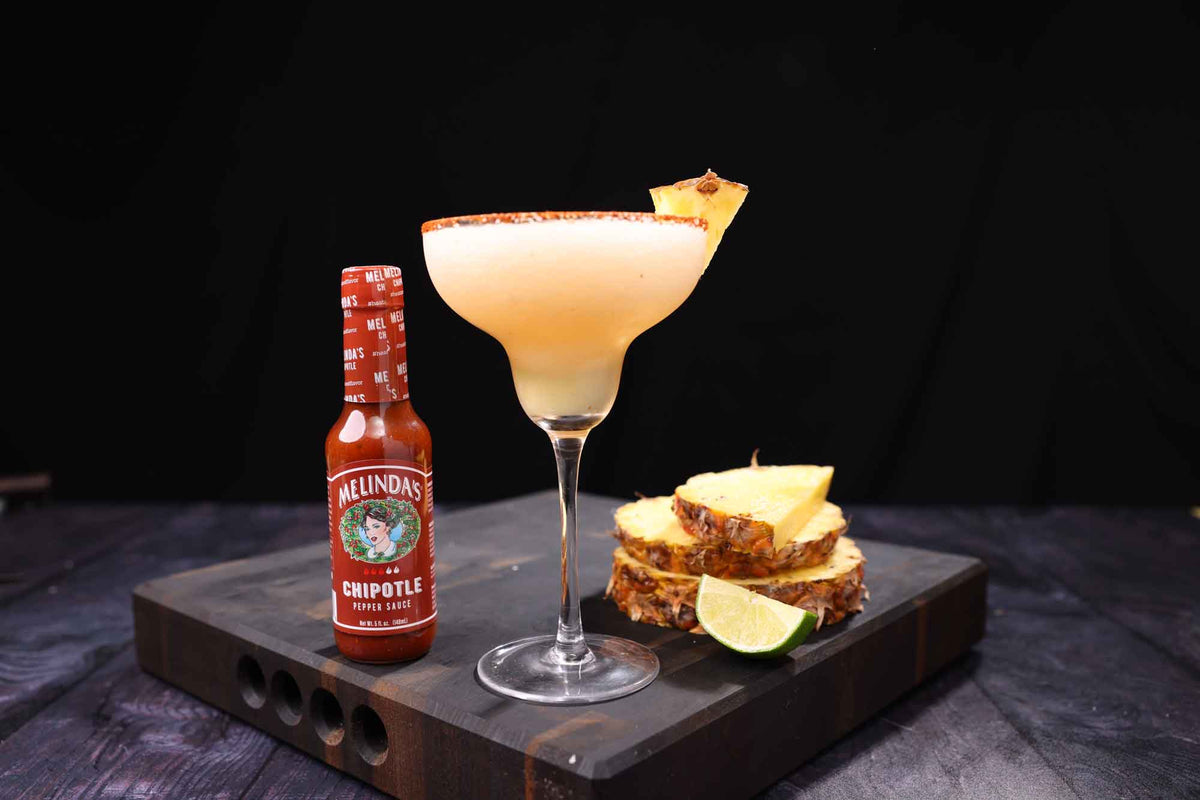 Creamy cocktail with pineapple garnish and spiced rim in margarita glass, with Melinda's Chipotle hot sauce bottle and...