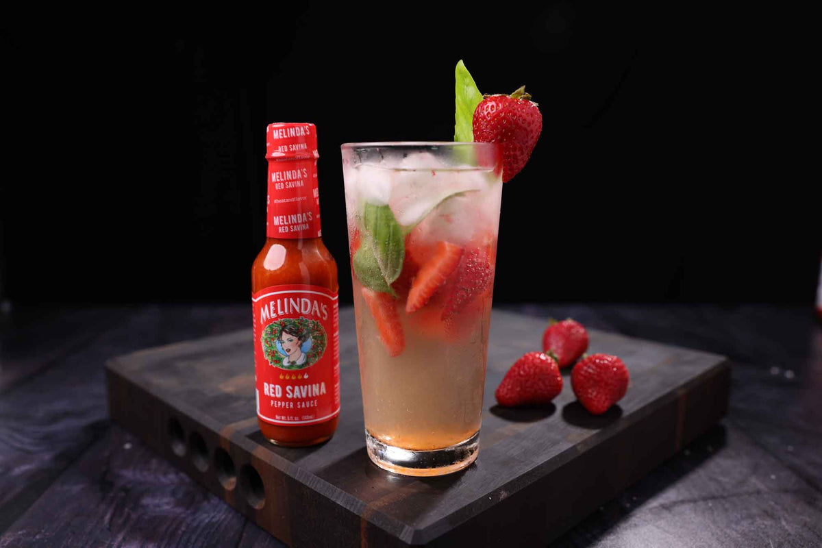 A tall glass cocktail with strawberries and mint sits next to a bottle of Melinda's Red Savina hot sauce on a wooden board.