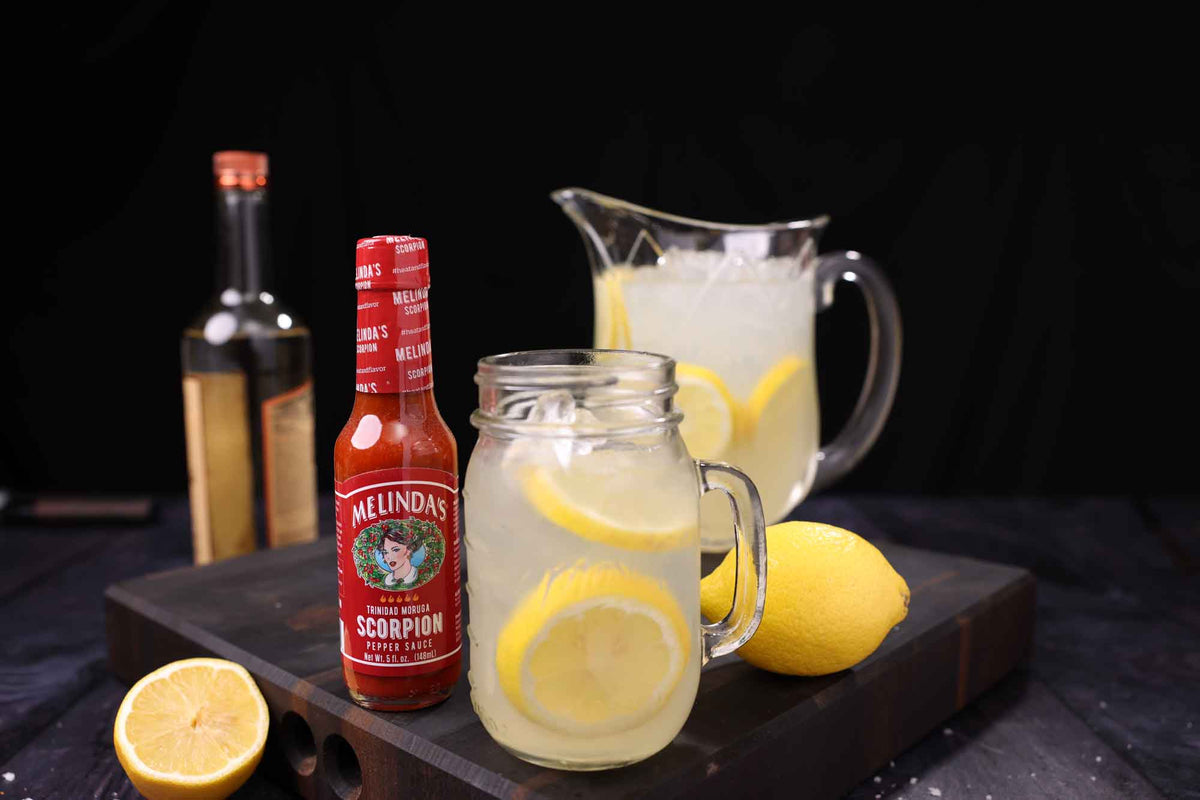 Bottle of Melinda's Trinidad Moruga Scorpion Pepper Sauce with lemonade pitcher, mason jar, and fresh lemons on wooden board