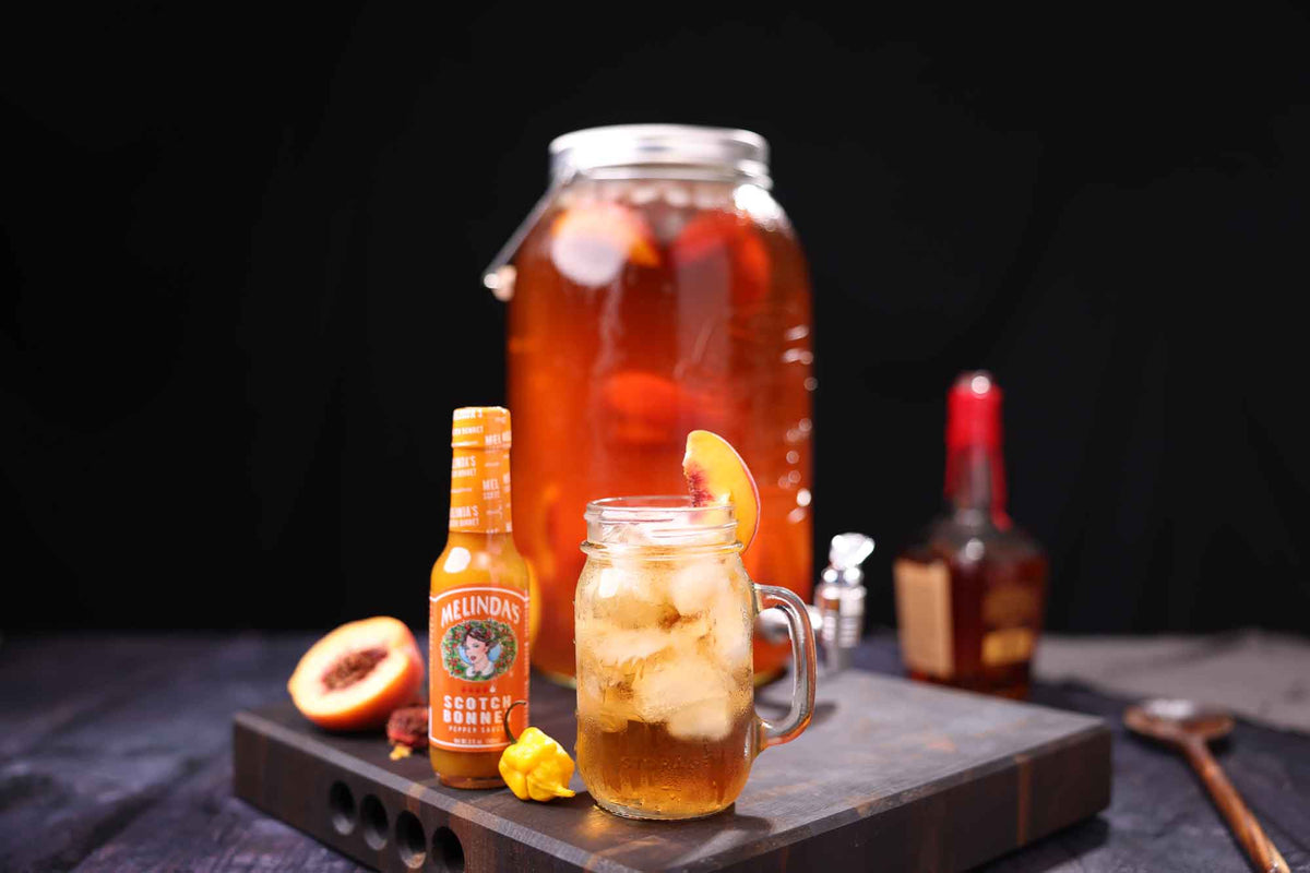 Melinda's Scotch Bonnet hot sauce bottle with cocktail ingredients including a mason jar drink, peach, and whiskey bottle