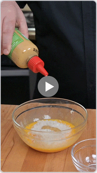 Person squeezing Melinda's hot sauce from bottle into glass bowl containing eggs on wooden surface