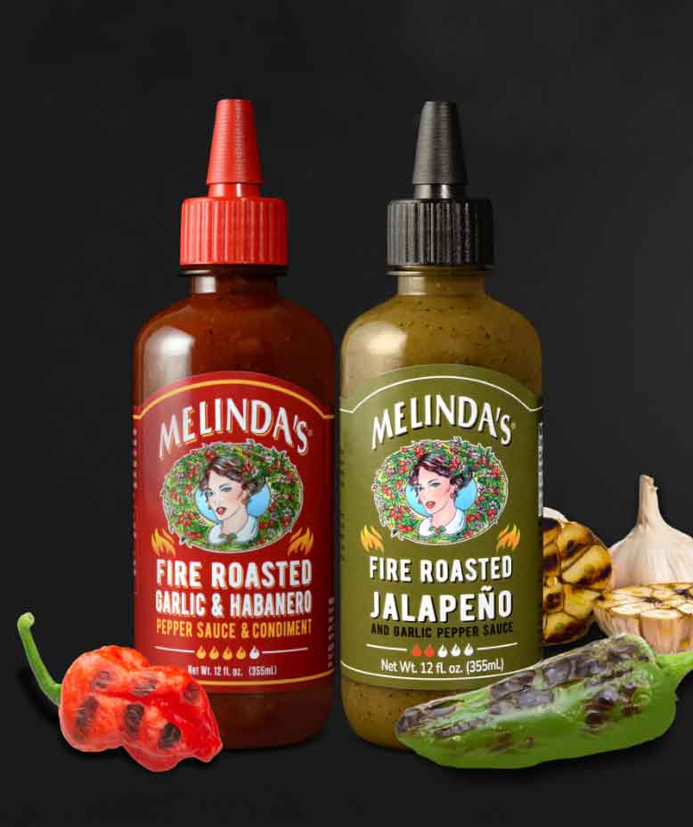 Two bottles of Melinda's hot sauce - Fire Roasted Garlic & Habanero and Fire Roasted Jalapeño - with fresh peppers and garlic