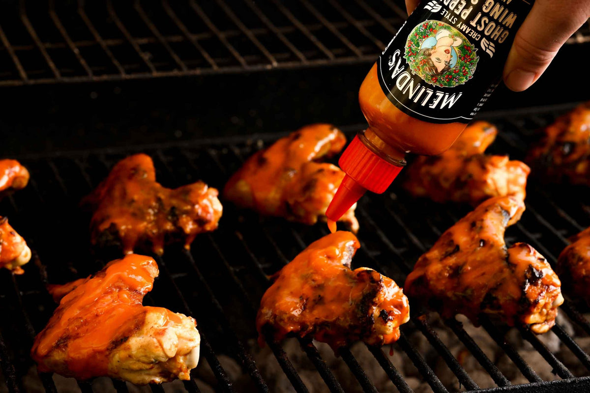 Chicken wings cooking on grill grates with hand pouring Melinda's Ghost Pepper hot sauce from bottle