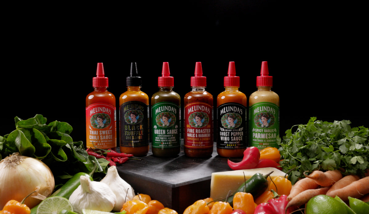 Six bottles of Melinda's hot sauce with red caps arranged on slate, surrounded by fresh vegetables and herbs on black...