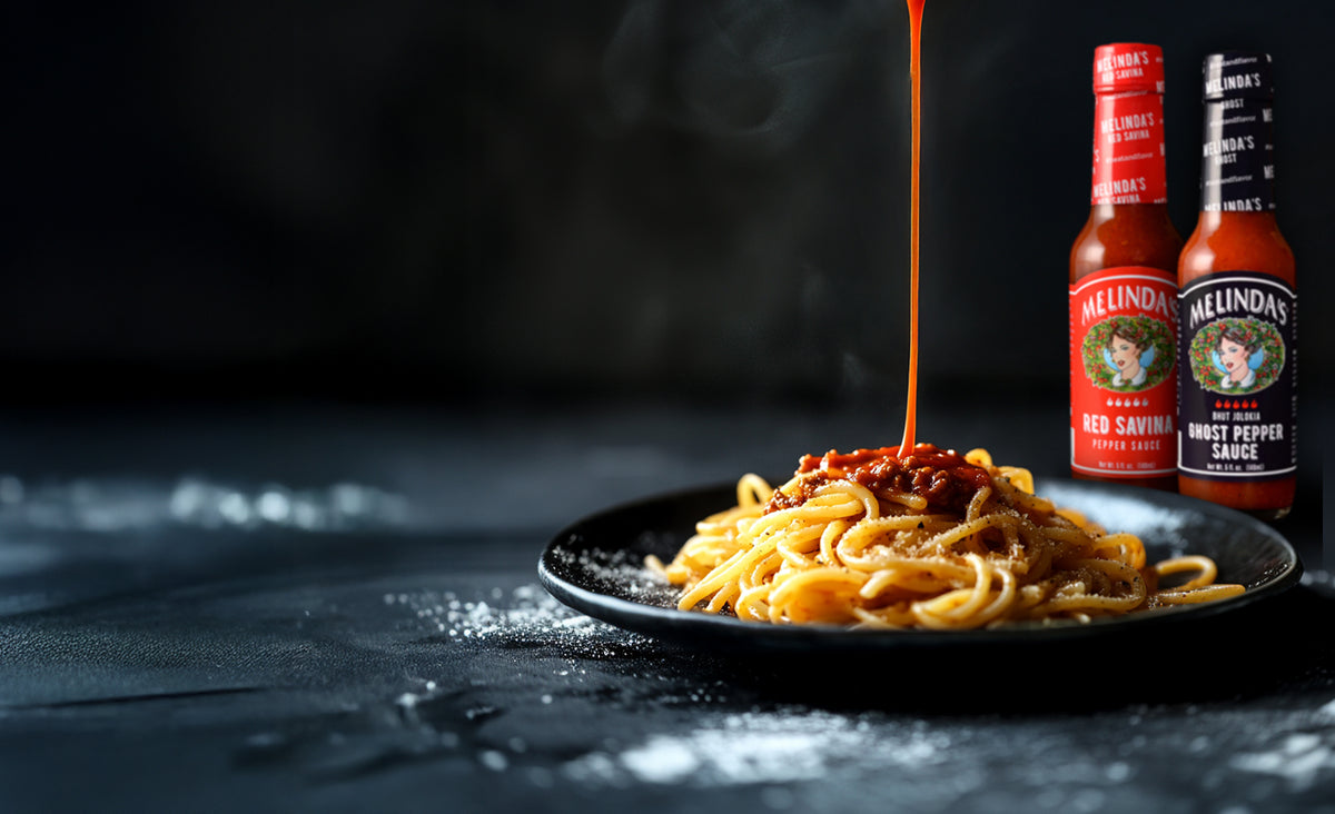 Plate of pasta with hot sauce being drizzled on top, accompanied by two bottles of Melinda's hot sauce on dark surface