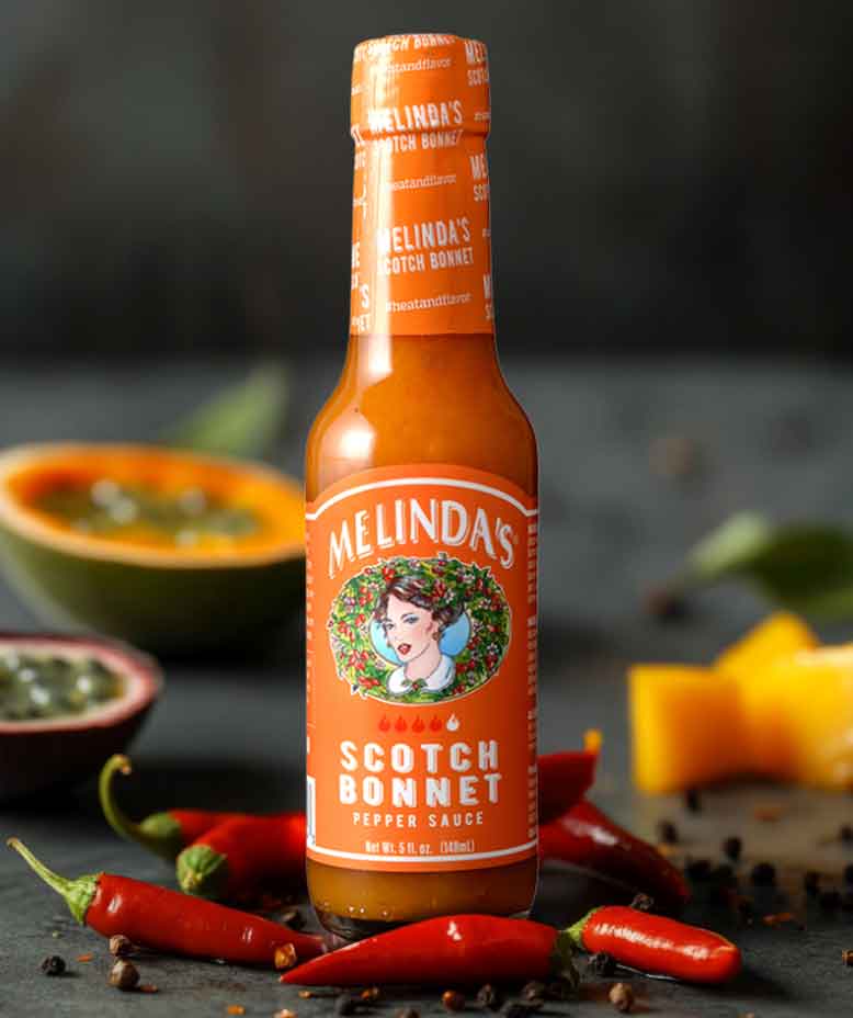 Melinda's Scotch Bonnet pepper sauce bottle surrounded by fresh red chilies and colorful vegetables on dark surface
