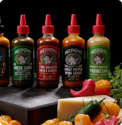 Four bottles of Melinda's hot sauce with red caps displayed on wooden surface with colorful peppers and herbs