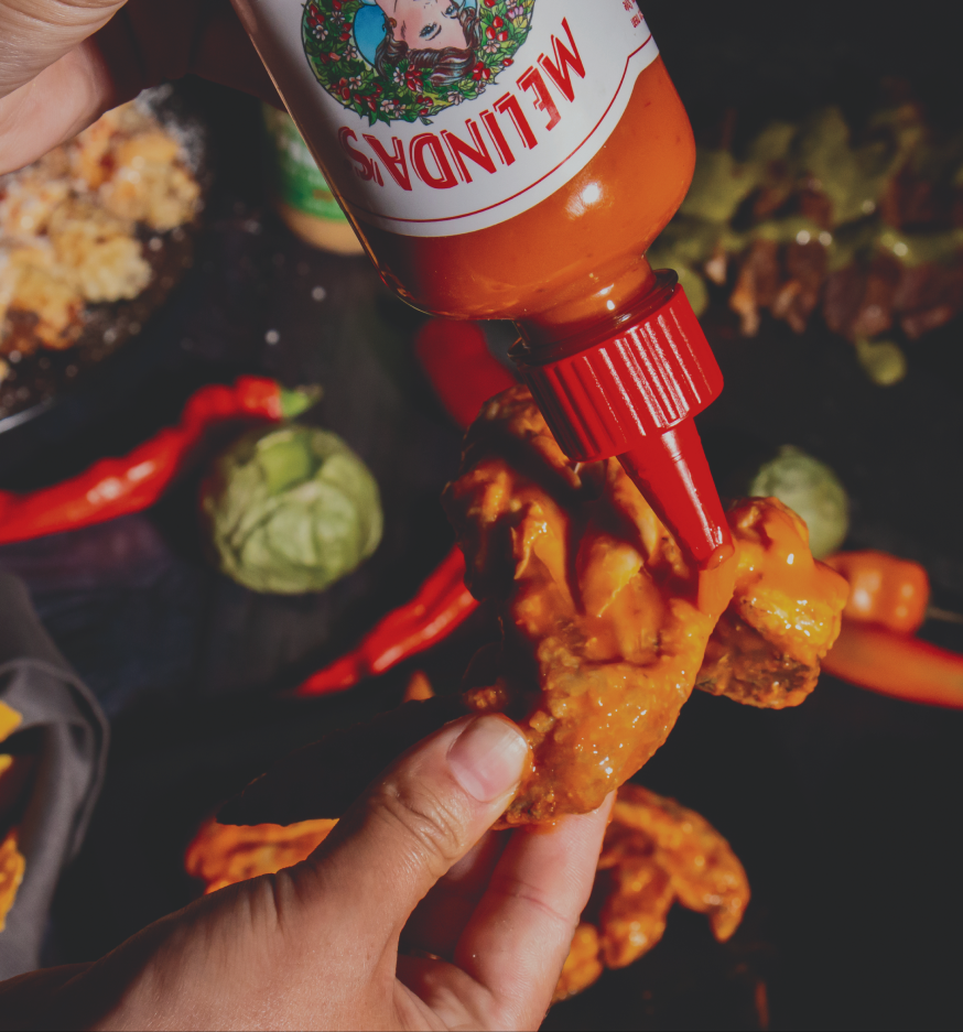 Hand holding Melinda's hot sauce bottle drizzling sauce onto crispy chicken wings on a dark plate with vegetables