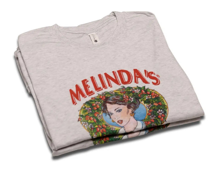Folded gray t-shirt with "MELINDA'S" logo in red text and colorful illustration of woman surrounded by floral wreath